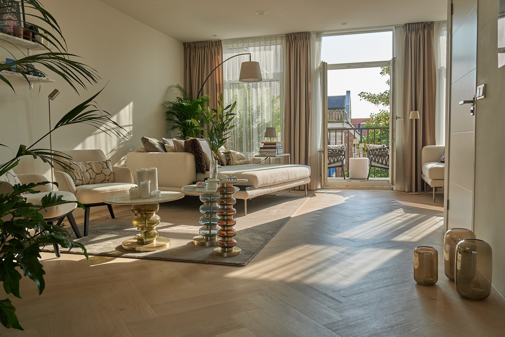 A living room from a full home renovation with chevron oak parquet, cream bouclé seating, potted palms and balcony doors open to the afternoon sun